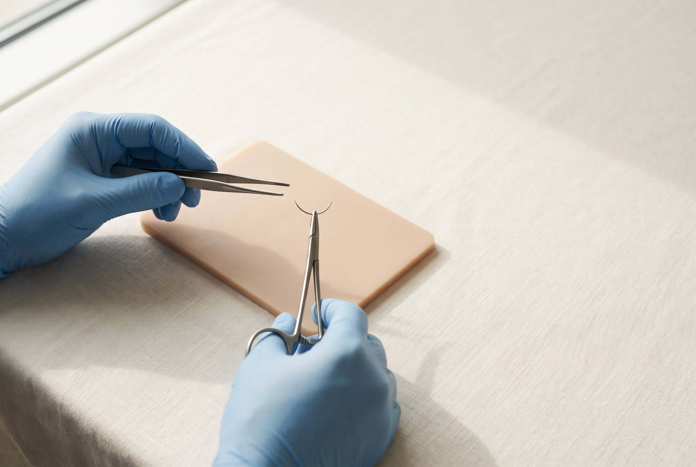 Gloved hands holding surgical forceps and a needle holder above a silicone skin simulation pad.